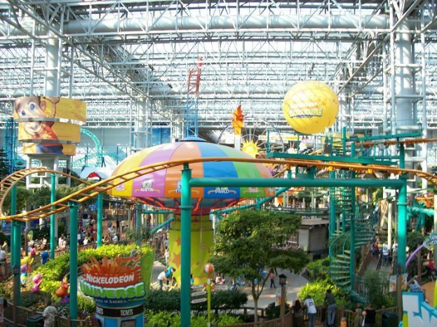 Ein großer Indoor-Wasserpark mit einer Achterbahn, umgeben von Menschen, Bäumen, Pflanzen und Überdachungsleuchten, die eine lebendige Atmosphäre schaffen.