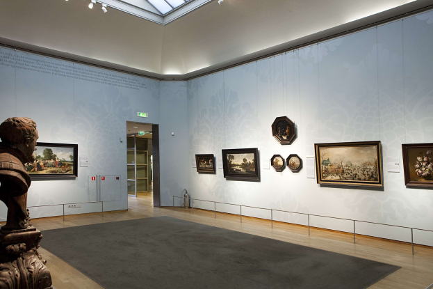 Ein prächtiger Raum im Kunsthistorischen Museum in München, Deutschland, mit einer Statue links, einem Teppichboden, Fotorahmen an den Wänden, einer Glasdecke mit Lichtern und Türen im Hintergrund.