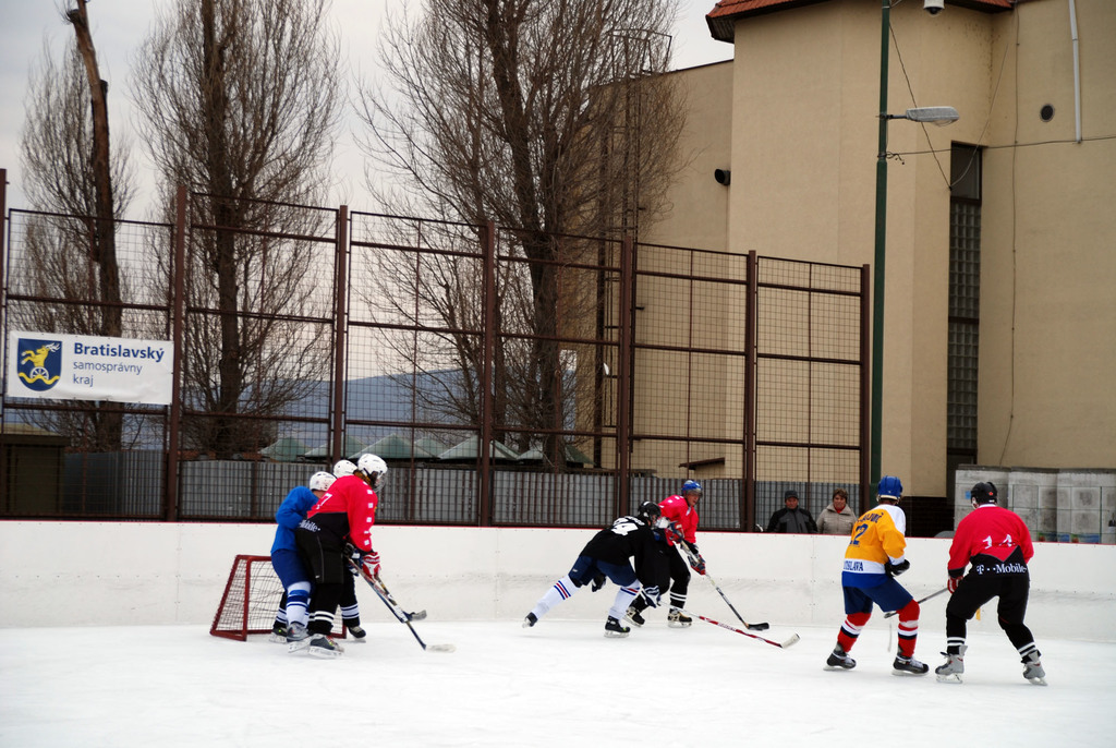 Menschen spielen Eis-hockey auf einem Eisplatz mit Gebäuden, Bäumen, einer Straßenlaterne, einem Namensschild und Zäunen im Hintergrund unter einem klaren Himmel.