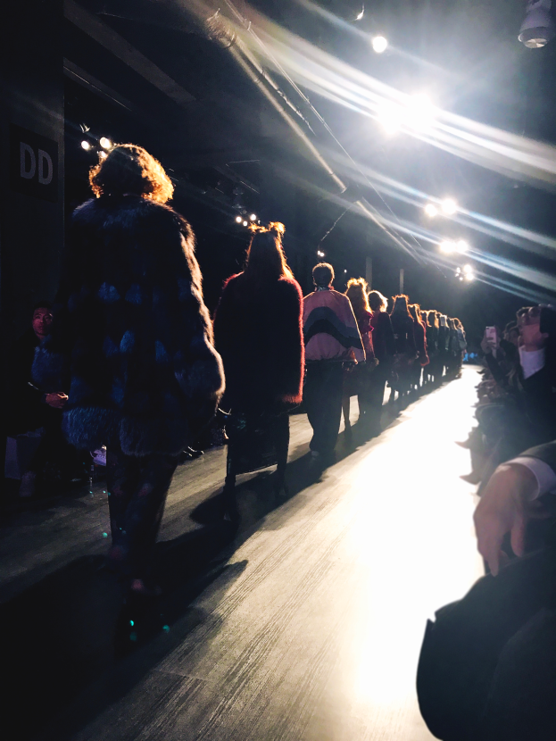 Eine Gruppe von Menschen läuft über den Laufsteg einer Fashion Show, beleuchtet von hellen Scheinwerfern, mit Zuschauern auf beiden Seiten.