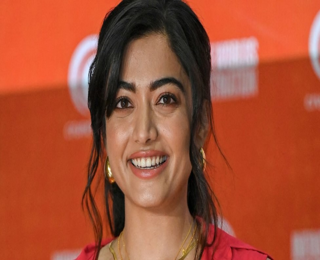Eine Frau in einem roten Kleid mit einem eleganten Dutt, schwarzen High Heels und dezentem Make-up lächelt in die Kamera und steht vor einem gelben Banner mit der Aufschrift 'Die besten Schönheits Looks von Sonam Kapoor beim Cannes Film Festival.'