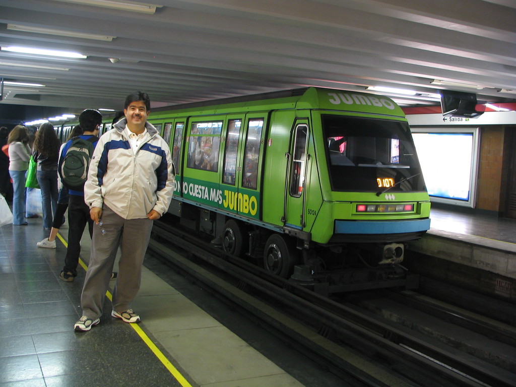 Ein Zug auf einem Bahngeleis mit Menschen auf der linken Seite und einem Dach am oberen Bildrand.