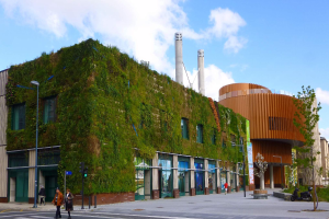 Ein großes Gebäude mit einer grünen Wand an der Seite, umgeben von Straßenlaternen, Verkehrspfosten, Verkehrsampeln, Kraftfahrzeugen, Fußgängern, Bäumen und einem bewölkten Himmel als Teil des Green Building Project.