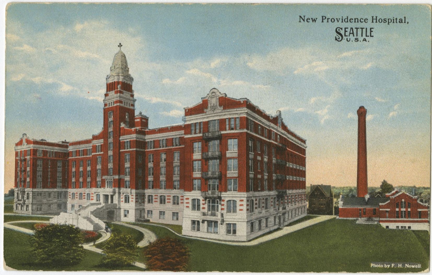 Eine Vintage-Postkarte, die das New Providence Hospital in Seattle, Washington zeigt, mit seinem mehrstöckigen Gebäude, Treppen, Geländern, Bäumen, Gras und einem bewölkten Himmel, einschließlich gedrucktem Text.