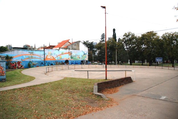 Eine Skate-Anlage mit einem Wandgemälde an einem Gebäude, umgeben von Gras, trockenen Blättern, Laternen, Geländern und einer bemalten Wand, vor einem Hintergrund aus Bäumen, Gebäuden, Fahrzeugen und Himmel.