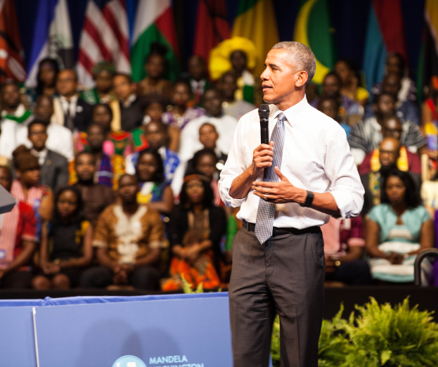 Präsident Obama spricht auf einer Wahlveranstaltung, hält ein Mikrofon, mit einer Menge von Menschen, einigen sitzend und einigen stehend, und einer Pflanze im Vordergrund, Flaggen und eine Tafel im Hintergrund.