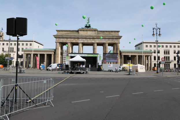 Brandenburger Tor in Berlin mit seinen Säulen und Statuen, umgeben von Gebäuden, Laternenmasten, Ampeln, Zelten, Fahrzeugen, Menschen, Fahrrädern und grünen Ballons vor einem bewölkten Himmel.