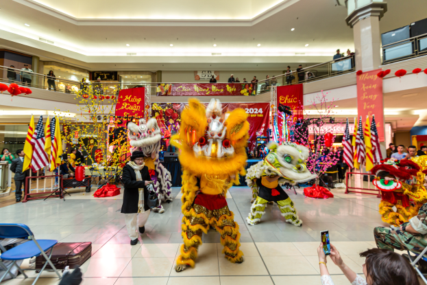 Ein lebendiges chinesisches Neujahrsfest in einem Einkaufszentrum mit tanzenden Menschen in Kostümen und anderen, die auf Stühlen sitzen und Mobiltelefone halten, umgeben von Fahnen, Bannern, Dekorationen und Lichtern.