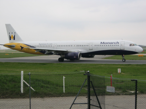 Ein Monarch Airlines Airbus A320-200 auf einer Rollbahn mit Gras, einem Zaun, Bäumen und einem klaren blauen Himmel im Hintergrund.