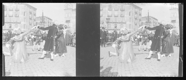 Ein Schwarz-Weiß-Foto einer Gruppe von Menschen, die auf der Straße tanzen, während eines Faschingsumzugs, gekleidet in Kostüme, mit Gebäuden, Straßenlaternen und -pfosten im Hintergrund bei klarem Himmel.