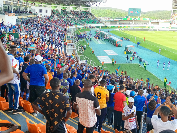 Eine große Menschenmenge versammelt sich vor einem eingezäunten Fußballfeld, wobei einige auf Stühlen sitzen und andere stehen, unter einem klaren blauen Himmel mit Hügeln im Hintergrund und warten gespannt auf das Spiel der afrikanischen Nationenmeisterschaft 2019.