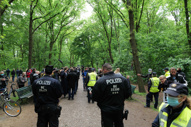 Eine Gruppe von Polizeibeamten steht vor einer Menschenmenge während einer Anti-Terror-Demonstration in Berlin, mit Fahrrädern und einer Bank im Vordergrund und Bäumen und Himmel im Hintergrund.