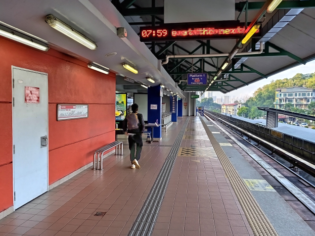 Ein Mann geht auf einem Bahnsteig neben einem stehenden Zug, der eine Tasche trägt, mit Bänken, Lampen, Informationsanzeigen und einer Tür in der Nähe, vor einem Hintergrund aus Bäumen, Gebäuden und Himmel.