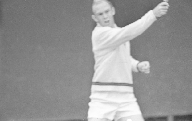 Ein Mann in weißer Tennisbekleidung steht auf einem Court und hält einen Schläger in der Hand, mit entschlossener Miene, umgeben von grünem Gras unter einem klaren blauen Himmel.