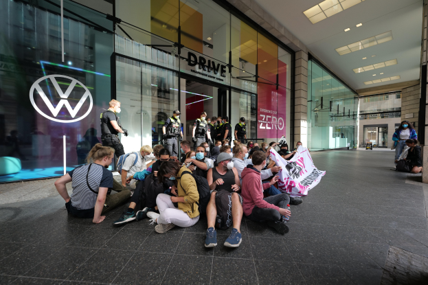 Eine Gruppe von Menschen sitzt vor einer Volkswagen-Filiale mit Glaswänden auf dem Boden und hält ein Transparent.