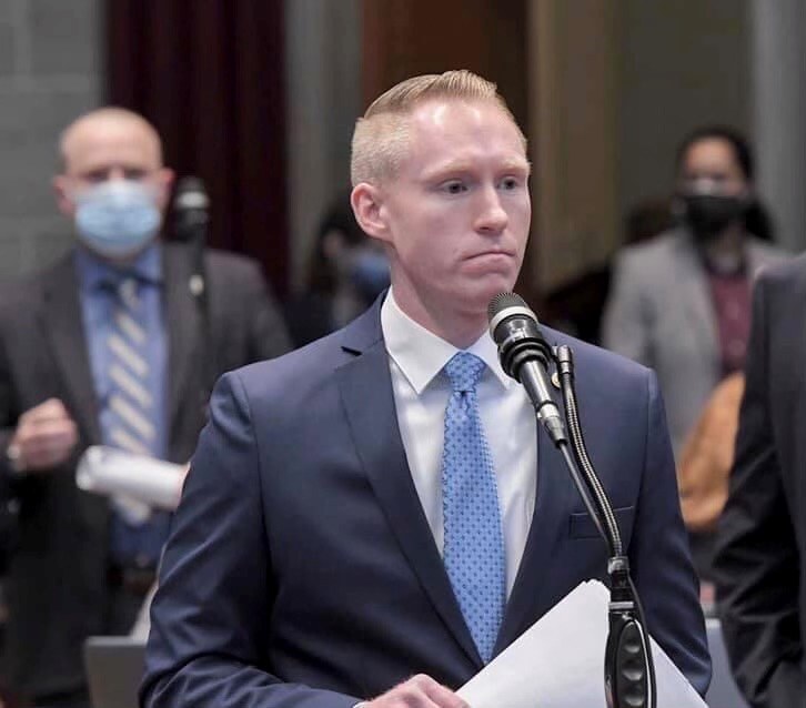 Ein Mann im Anzug spricht an einem Podium mit Papieren vor einem Mikrofon, während einige Menschen hinter ihm in der Nähe einer Vorhangwand stehen.