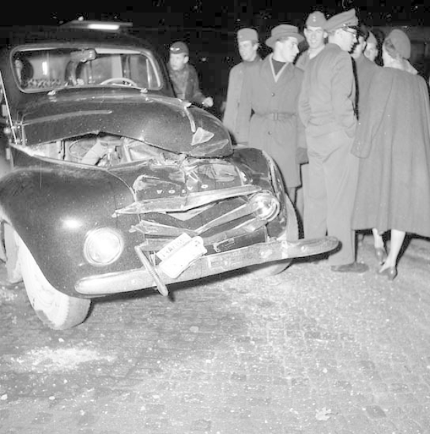 Schwarz-weißes Bild eines schwer beschädigten Autos am Straßenrand mit einer Gruppe von Menschen, die es inspizieren.