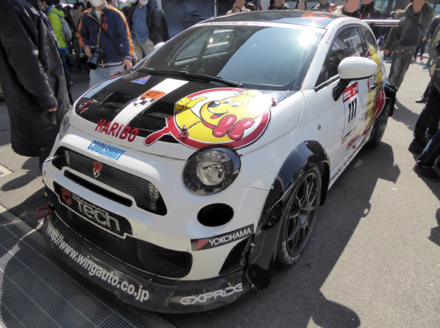 Ein kleiner weißer Wagen vor einer Menschenmenge geparkt, einige tragen Masken und halten Kameras, mit Gebäuden und einem klaren blauen Himmel im Hintergrund.