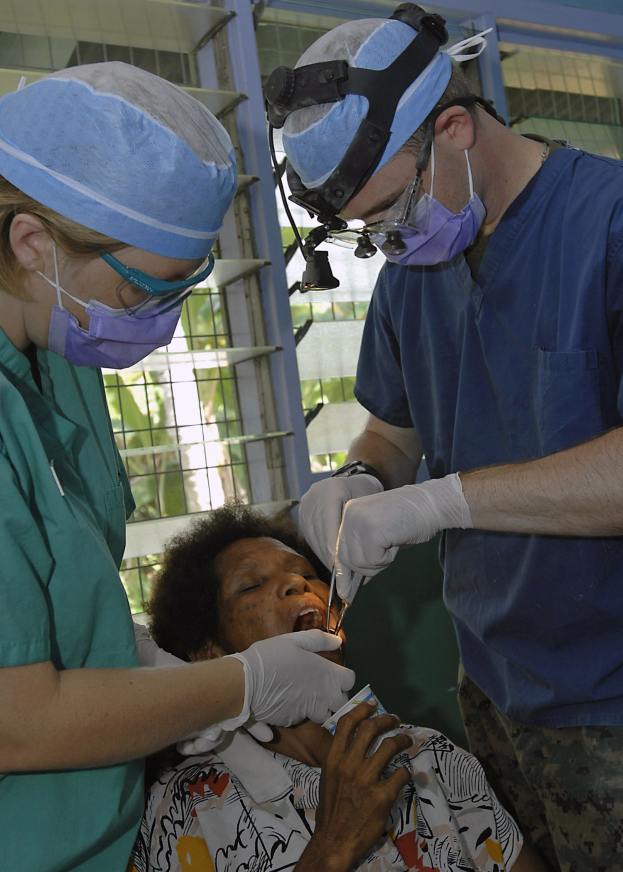 Zwei Chirurgen in Masken, Kappen und Handschuhen führen eine Gesichtsoperation an einer Frau durch, die auf einem Bett liegt, mit einem Fenster und Bäumen im Hintergrund.