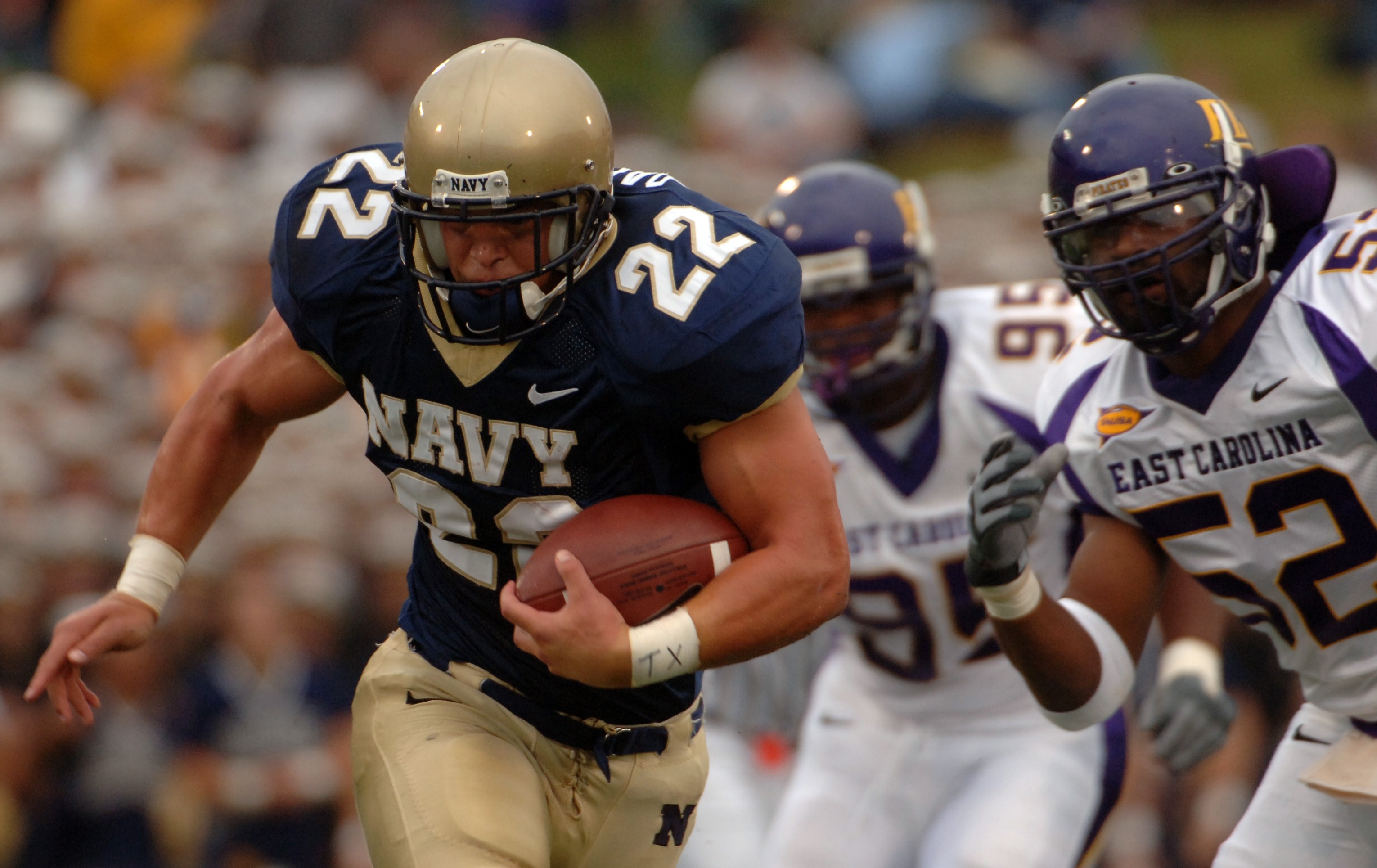 Ein Football-Spieler in einem marineblauen Trikot und Helm rennt mit dem Ball während eines Spiels, mit einem unscharfen Hintergrund, der schnelle Bewegung andeutet.