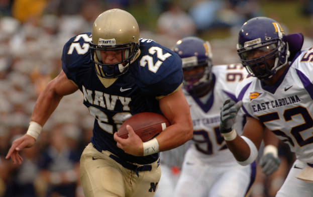 Ein Football-Spieler in einem marineblauen Trikot und Helm rennt mit dem Ball während eines Spiels, mit einem unscharfen Hintergrund, der schnelle Bewegung andeutet.