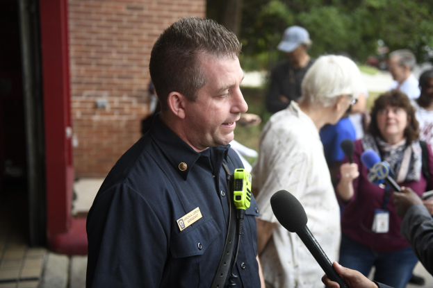 Ein Polizist in Uniform, der mit einer Gruppe von Menschen spricht, die Mikrofone halten, vor einem Hintergrund aus Bäumen und einem Gebäude.