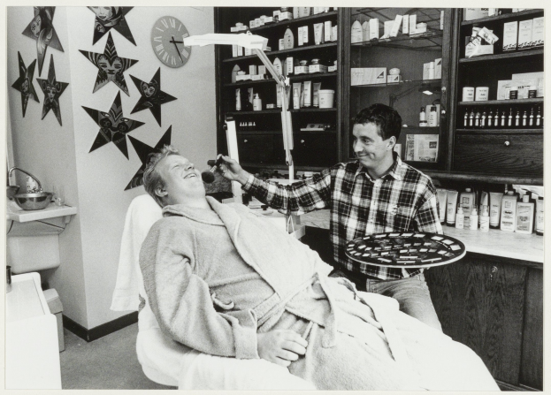 Ein Mann lächelt im Zahnarztstuhl, während ein anderer Mann neben ihm steht und ein Tablett hält, mit einem Waschbecken, einer Uhr an der Wand und Regalen mit Flaschen im Hintergrund.