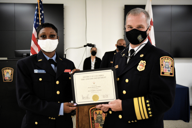 Zwei Männer in Uniform und Masken stehen nebeneinander und halten ein Zertifikat, mit einem Podium mit Mikrofon dahinter, Flaggen, einer Wand, einem Tisch mit einem Monitor links und einem Tisch mit blauer Decke rechts.