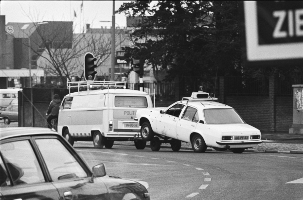 Schwarz-weiß-Foto eines Wagens, der von einem Polizeiwagen auf einer Stadtstraße geschleppt wird, mit anderen Fahrzeugen, einem Fußgänger, Ampeln, Masten, Bäumen, Gebäuden und einem Zaun im Hintergrund.