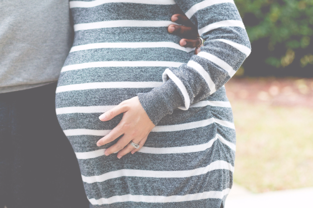 Schwangere Paare stehen zusammen mit den Händen auf dem Bauch, tragen Schwangerschaftskleidung in einer holzigen Outdoor-Umgebung.