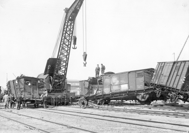 Schwarzes und weißes Bild von einer Zugkatastrophe mit einem Kran, der ein Waggon auf die Schienen hebt, mehrere Personen in der Nähe und im Hintergrund Gebäude unter einem klaren Himmel.