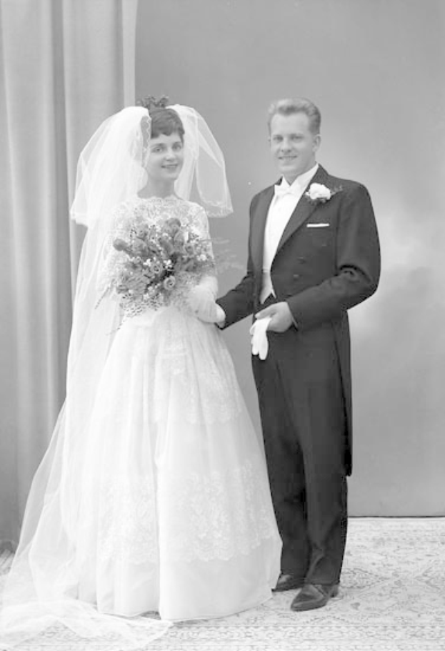 Ein Schwarz-Weiß-Foto eines Mannes und einer Frau, die Hand in Hand auf ihrer Hochzeit stehen, wobei die Frau ein weißes Kleid trägt und einen Strauß hält und der Mann strahlt, mit einem Vorhang und einer Wand im Hintergrund.