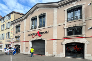 Eine Gruppe von Menschen mit Fahnen geht vor einem Gebäude mit einer Tafel, die "Montbron" lautet, einer Straßenlaterne und einer Fahne mit Bildern und Text unter einem bewölkten Himmel.