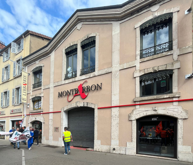 Eine Gruppe von Menschen mit Fahnen geht vor einem Gebäude mit einer Tafel, die "Montbron" lautet, einer Straßenlaterne und einer Fahne mit Bildern und Text unter einem bewölkten Himmel.