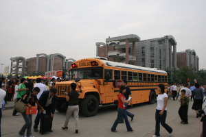 Eine Gruppe von Menschen, einige tragen Taschen, geht vor einem Schulbus auf einer Straße, die von Gebäuden, Bäumen und Laternen gesäumt ist, unter einem klaren blauen Himmel.