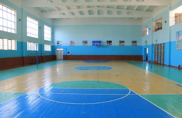 Große Turnhalle mit blau-weißem Boden, Basketballfeldmarkierungen, einem Netz an Pfosten, einer Tür, Fenstern, Wandplakaten, einem Geländer und einem Dach.