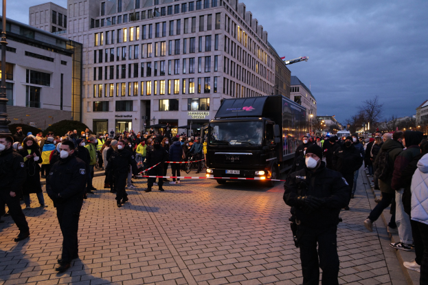 Eine Gruppe von Menschen steht in der Nähe eines Lastwagens auf einer Straße mit Gebäuden, Bäumen und Laternen im Hintergrund; einige tragen Mützen und Masken und ein Band an einem Pfahl ist im Vordergrund zu sehen.