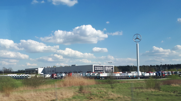 Ein Mercedes-Benz-Werk in Deutschland mit Fahrzeugen auf der Strasse, umgeben von Grünflächen und Gebäuden unter einem bewölkten Himmel.