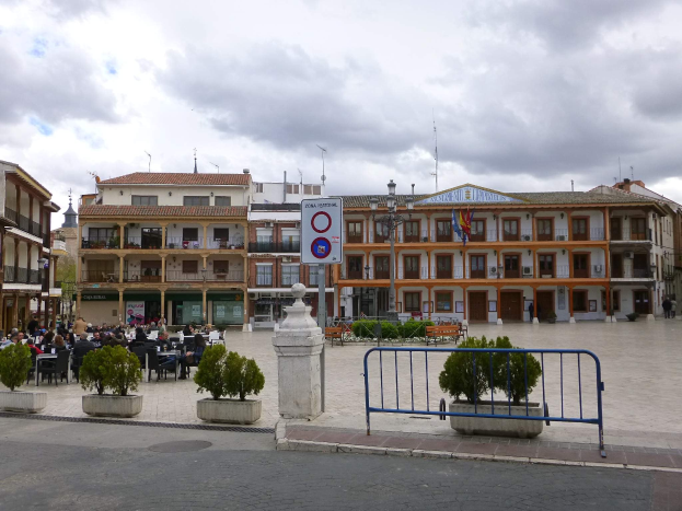 Ein belebter Stadtplatz mit Menschen auf Stühlen sitzend und stehend, umgeben von Topfpflanzen, Metallabsperrungen, Schildern an Pfosten, Fahnen, Gebäuden und einem bewölkten Himmel.