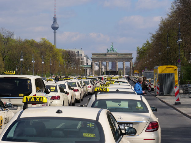 Eine belebte Straße in Berlin mit zahlreichen geparkten Taxis, Fußgängern auf dem Gehweg, gesäumt von Laternenmasten und Bäumen, Gebäuden im Hintergrund, einem fernen Bogen mit Statuen und Turm und einem bewölkten Himmel.