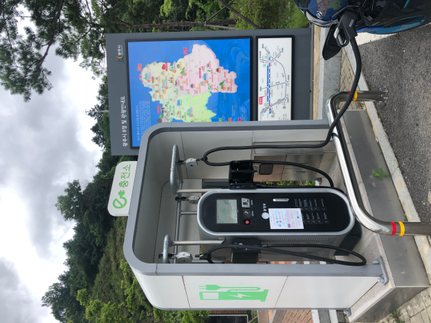 Eine Ladestation für Elektrofahrzeuge am Straßenrand mit einem Motorrad daneben, ein digitales Anzeigetafel, die anzeigt, dass es eine E-Bike-Ladestation ist, mit Bäumen und einem klaren blauen Himmel im Hintergrund.