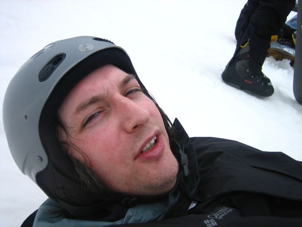 Ein Mann in Pullover und Helm steht im Schnee, mit den Beinen und Skiern einer anderen Person in der oberen rechten Ecke.