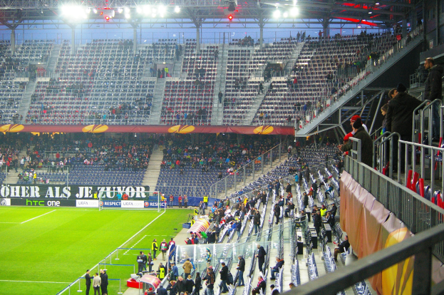 Großes Stadion voller Zuschauer bei einem Fußballspiel mit sitzenden und stehenden Personen, einer Absperrung, einem Banner auf der rechten Seite und einem leeren Abschnitt im Hintergrund mit Deckenbeleuchtung.
