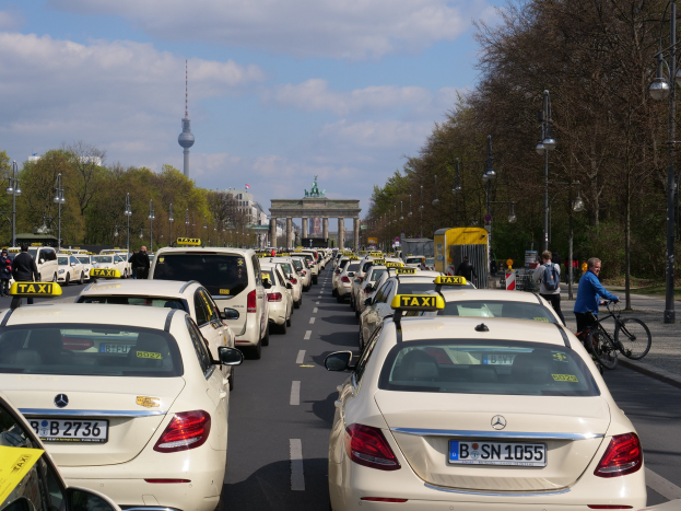 Lange Reihe von Taxis entlang einer belebten Straße in Berlin, Deutschland, mit Fahrradfahrern und Fußgängern auf dem Gehweg, flankiert von Laternen und Bäumen und Gebäuden, einem Bogen und einem Turm im bewölkten Hintergrund.