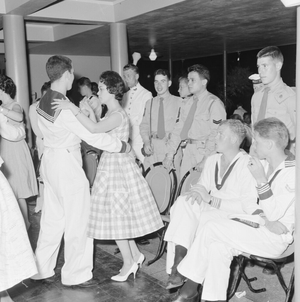 Schwarz-weiß-Foto einer gemischten Geschlechtergruppe in Militäruniformen, die auf einem Tanzboden tanzen und sitzen, mit Säulen und Beleuchtung im Hintergrund.