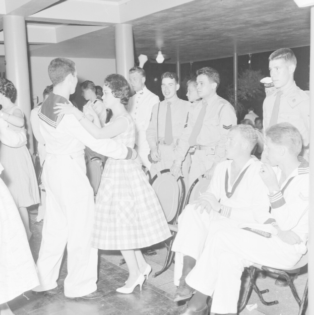 Schwarz-weiß-Foto einer gemischten Geschlechtergruppe in Militäruniformen, die auf einem Tanzboden tanzen und sitzen, mit Säulen und Beleuchtung im Hintergrund.