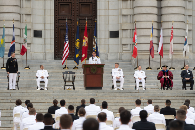 Gruppe von Menschen in weißen Uniformen sitzt auf den Stufen eines Gebäudes mit einem Podium, das von Fahnen flankiert ist, vor einem großen Gebäude mit einer Tür während einer offiziellen Zeremonie.