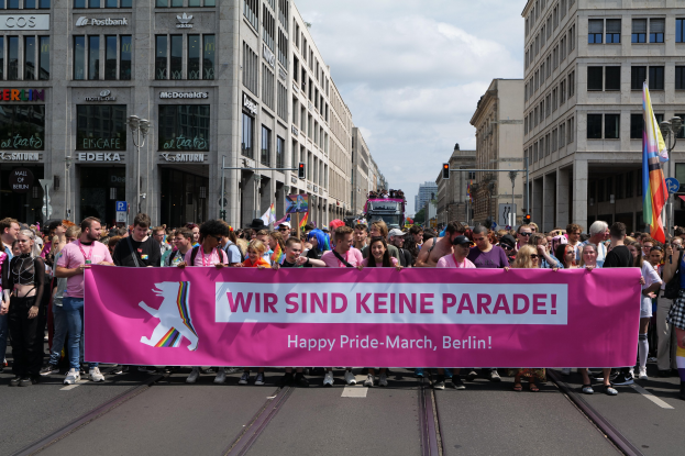 Eine Gruppe von Menschen marschiert auf einer Straße in Berlin, Deutschland, mit einem pinken Banner, auf dem "Happy Pride March" steht, während Gebäude, Laternenpfähle und Verkehrszeichen die Straße säumen.