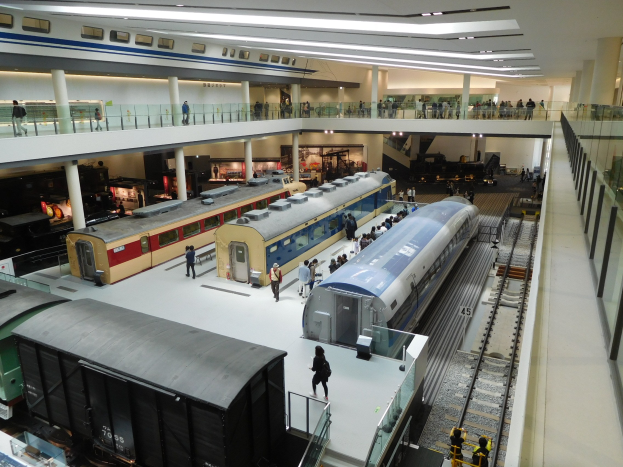 Ein großer Museumsraum mit verschiedenen Zügen in unterschiedlichen Gröüen und Farben, bei denen Menschen die Ausstellungsstücke und architektonische Elemente wie Geländer, Säulen und Deckenleuchten betrachten.