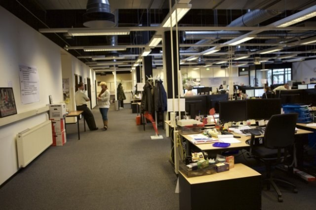 Großes Großraumbüro mit Menschen an Schreibtischen, die mit Computern, Monitoren, Tastaturen und Papieren ausgestattet sind, umgeben von Stühlen, Büchern, Kisten und hängenden Kleidungsstücken, mit an der Wand montierten Tafeln mit Text und Bildern, Deckenleuchten und Glasfenstern im Hintergrund.